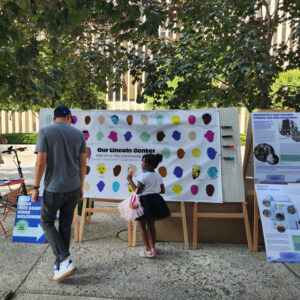 A man and a child view the community mural. They are on a sidewalk; the exhibit invites them to draw their own portrait.